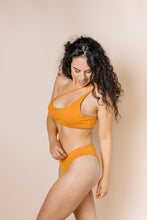 Load image into Gallery viewer, Girl in burnt orange bikini set. Top is asymmetrical with the straps coming up to the left shoulder. Side view
